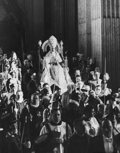 Pope Pius XII wearing his ecclesiastical garb, incl. decorative miter, & being carried on his papal throne while Swiss & Noble guardsmen join entourage, at St. Peter's. - 1940